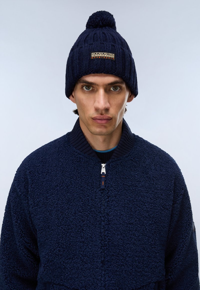 Jonge man met een donkerblauwe gebreide muts met pompon en een gestructureerd donkerblauw zip-up jack op een lichtgrijze achtergrond.