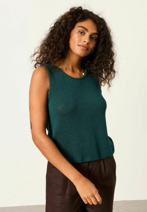 Mladá žena kučeravými vlasmi, oblečená v tmavozelenom tielku bez rukávov a tmavých nohaviciach, stojaca na jednoduchom svetlom pozadí.