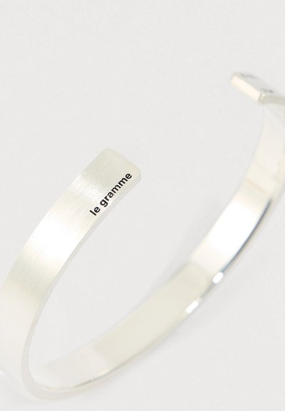 Srebrna kovinska zapestnica s brušenim zaključkom in vgraviranim "le gramme" blizu enega konca na belem ozadju.