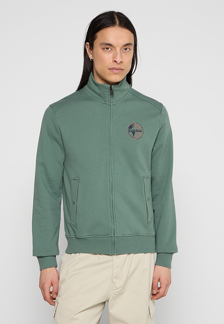 Felpa verde con zip e colletto alto, dotata di due tasche frontali e un logo circolare sul petto, realizzata in un tessuto morbido.