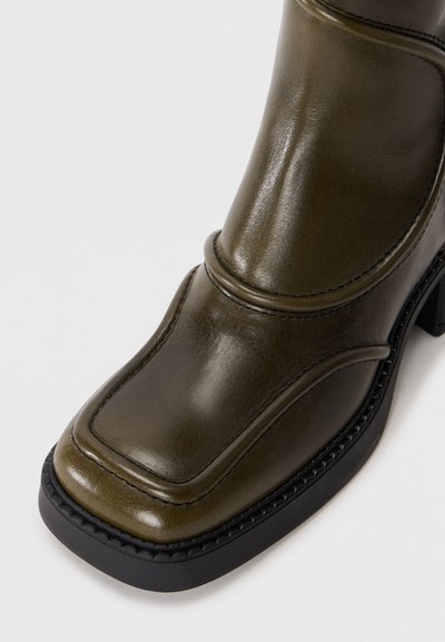 Stivaletto verde in pelle fino alla caviglia con punta quadrata, cuciture decorative e suola nera spessa. Texture liscia con leggera lucentezza.