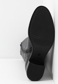 Botte en cuir noir texturé avec fermeture éclair latérale, présentée avec la semelle vers le haut montrant une semelle extérieure en caoutchouc à motif et un talon bloc sur fond blanc.