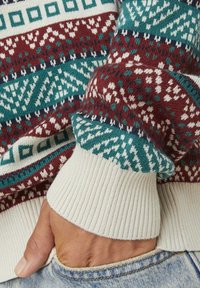 Gebreide sweater met een bordeauxrood, teal en witte geometrische patroon. Geribde manchetten in een crème kleur. Zachte textuur. Hand in de denimzak.