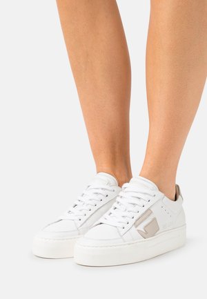 White sneakers with a textured leather upper, raised rubber sole, and beige accents. Features lace-up design and perforated detailing.