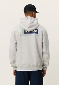 Ανοιχτό γκρι φούτερ με κουκούλα και μεγάλο μπλε-πράσινο γραφικό στο πίσω μέρος που γράφει "LES DEUX SPORTING GOODS." Διαθέτει ρυθμιζόμενη κουκούλα με κορδόνια και ριμπ μανσέτες.