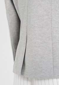 Šedý textilní vrch s texturovaným povrchem, s bočním rozparkem a rovným střihem. Výrazné švy a okraje jsou čisté a minimalistické.