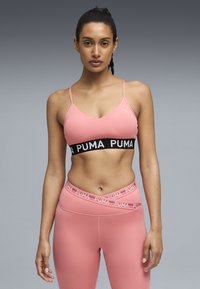 Γυναίκα με ροζ αθλητικό σουτιέν και κολάν με σήμα Puma, όρθια μπροστά σε απλό γκρι φόντο, κοιτάζοντας προς τα εμπρός με ουδέτερη έκφραση.