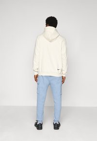 Man wearing a cream hoodie and light blue cargo sweatpants, standing facing away on a plain light gray background.