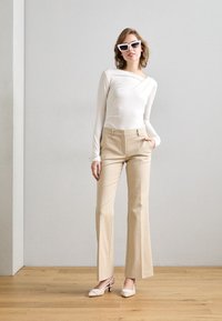 Blusa blanca de manga larga con diseño drapeado, pantalones beige de campana y zapatos de tacón bicolores. Los accesorios incluyen gafas de sol blancas con forma angular.