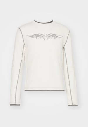 Chemise blanche à manches longues avec un motif tribal gris sur la poitrine et des coutures grises visibles le long des manches et des ourlets.