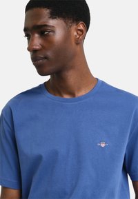 Blå bomulls-T-shirt med rund halsringning, korta ärmar och en liten broderad logo på bröstet. Slät textur och avslappnad passform.