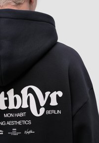 Černá mikina s kapucí vyrobená z silného materiálu. Na zadní straně je bílý grafický potisk s textem a logem. Kapuce je podšívkovaná hladkým materiálem.