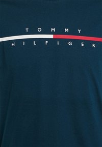 Tmavomodrá látka s textem "TOMMY HILFIGER" rozděleným vodorovnými bílými a červenými obdélníkovými pruhy uprostřed.