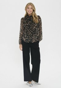 Schwarze und beige Bluse im Zebra-Print mit gerafftem Ausschnitt; kombiniert mit weit geschnittenen schwarzen Hosen und metallisch silbernen Schuhen.