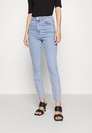 Kvinde iført lyseblå højtaljede skinny jeans, sort kortærmet top og sorte åbne høje hæle sandaler, stående mod en ensfarvet baggrund.