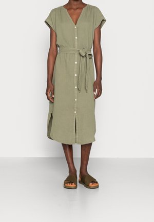 Vrouw draagt een knielange olijfgroene jurk met knopen aan de voorkant, een ceintuur in de taille, en bruine open-toe slip-on sandalen.