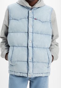 Smanicato imbottito in denim azzurro chiaro con bottoni a pressione, design trapuntato, due tasche anteriori e orlo arrotondato, indossato sopra una felpa grigia.