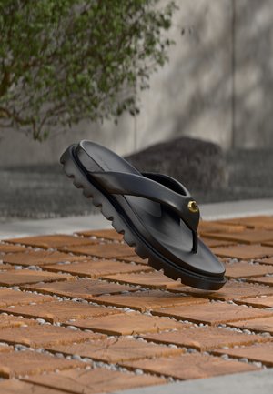 Sandale noire à tongs avec une semelle épaisse et robuste et un accent doré, flottant au-dessus d'une surface extérieure en briques texturées et gravier.