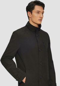 Cappotto nero con collo alto, bottoni frontali e tasca sul petto con zip. Realizzato in un tessuto liscio e opaco con tasche laterali.