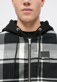Tânăr îmbrăcat cu un hanorac cu fermoar în carouri negre, albe și gri, cu glugă neagră și patch cu logo-ul Karl Lagerfeld Jeans pe piept.