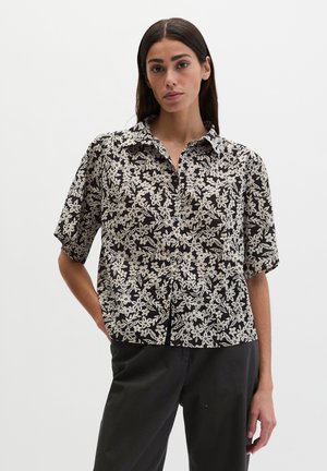 Kobieta w czarnej bluzce z krótkim rękawem w biały kwiatowy wzór i czarnych spodniach, stoi z jedną ręką w kieszeni na białym tle.