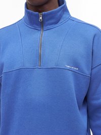 Pull bleu avec fermeture éclair quart et col montant, arborant le logo "Hollister" sur la poitrine, porté par une personne.