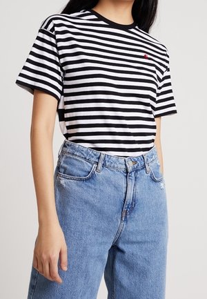 Osoba ubrana w czarno-biały t-shirt w poziome paski z małym czerwonym logo oraz wysokim stanem jasnoniebieskich dżinsów.