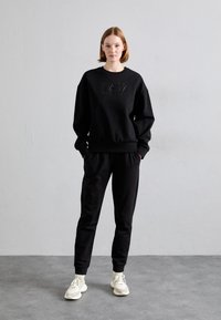 Μαύρη μπλούζα και παντελόνι από μαλακό ύφασμα. Η μπλούζα διαθέτει ανάγλυφο λογότυπο μπροστά. Το μοντέλο φοράει ανοιχτόχρωμα αθλητικά παπούτσια.