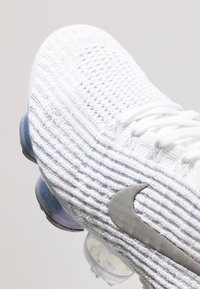 Bílé sportovní boty s texturovaným, pleteným svrškem. Vyznačují se šedým logem Nike a průhledným vzduchovým polstrováním na podrážce. Šněrovací design.