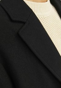 Abrigo de lana negro con solapas afiladas, combinado con un suéter de punto crema texturizado. Primer plano de los detalles de la tela y la costura.