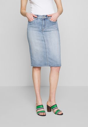Kvinde iført lysblåt knælangt denims nederdel, hvid top og grøn-sorte stribede hælede sandaler, står med hænder i nederdelslommerne.