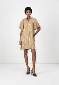Abito beige a maniche corte con un delicato motivo a catena e una chiusura con bottoni, abbinato a tacchi neri. Tessuto morbido e vestibilità rilassata.