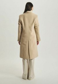 Manteau en laine beige avec une fente au dos, design ajusté et manches longues. La personne le porte avec un pantalon brillant et ajusté.