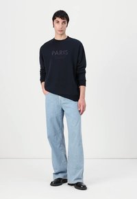 Felpa navy con "PARIS" stampato in grigio scuro, abbinata a jeans svasati blu chiaro e scarpe nere, su uno sfondo neutro.