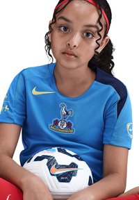Maillot de football bleu avec logo et accents jaunes, tissu texturé, associé à un ballon de football blanc et bleu marine avec un motif bleu.
