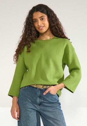 Femme aux longs cheveux bouclés portant un haut vert à manches longues et un jean bleu avec une ceinture beige, debout avec une main dans la poche.
