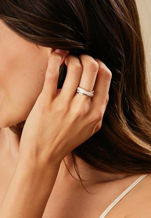 Silberner Ring mit ineinander verwobenen Bändern, polierter Oberfläche und subtilen glitzernden Akzenten, auf einer Hand präsentiert, die das Haar berührt.