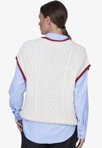 Witte gebreide vest met kabelpatronen en rode accenten bij de halslijn en armsgaten, gedragen over een lichtblauw shirt.