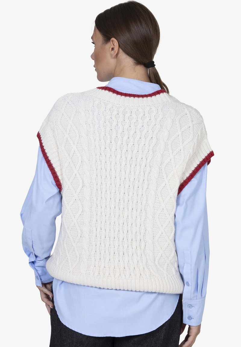 Witte gebreide vest met kabelpatronen en rode accenten bij de halslijn en armsgaten, gedragen over een lichtblauw shirt.