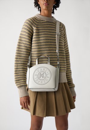 Un sac à main en cuir blanc avec des détails de logo perforés, une forme structurée, des double poignées supérieures et une bandoulière réglable. Porté avec un pull rayé.