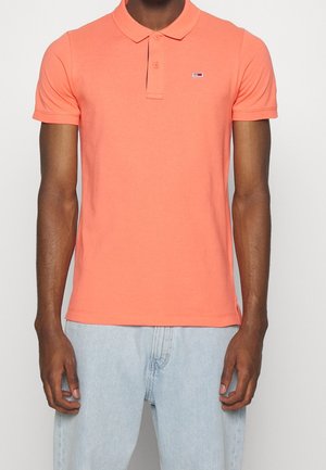 Homme portant un polo corail à manches courtes avec un petit logo sur la poitrine et un jean bleu clair, debout devant un fond uni.