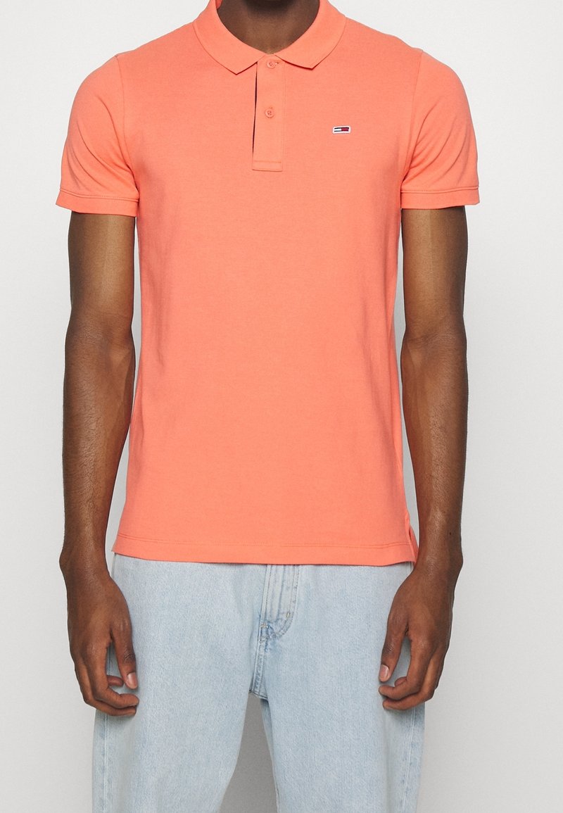 Homme portant un polo corail à manches courtes avec un petit logo sur la poitrine et un jean bleu clair, debout devant un fond uni.