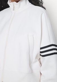 Valkoinen vetoketjullinen takki, jossa on korkea kaulus, teksturoitu pinta ja musta kolmiraitainen yksityiskohta vasemmassa hihassa. Sisältää sivutaskun.