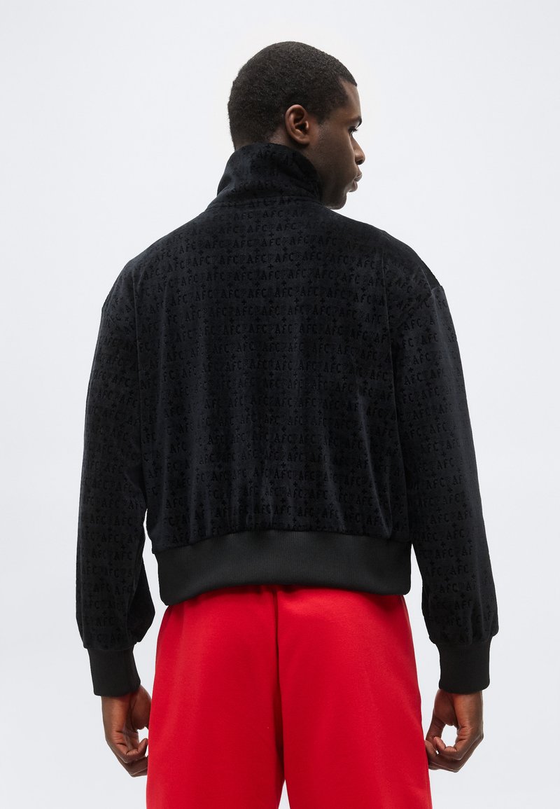Man draagt een zwarte getextureerde jas met een herhaald "AFC"-patroon, gecombineerd met felrode broek, kijkt weg tegen een effen achtergrond.