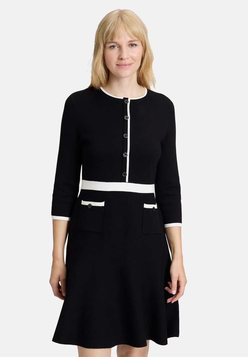 Robe noire avec un buste ajusté et une jupe évasée, présentant des accents blancs au niveau de l'encolure, des poignets et de la taille, ainsi que deux poches avant.