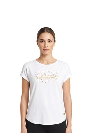 Une femme porte un T-shirt blanc orné d’un logo doré en écriture cursive et de détails cloutés, associé à un pantalon noir sur un fond blanc uni.