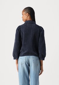 Navy corduroy blouse with puffed sleeves and buttoned cuffs. The back features a rounded hem and a classic collar design.