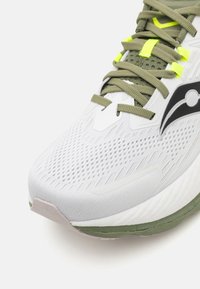 Hvit atletisk sko med teksturert mesh-overdel, grønne snøringer, svarte detaljer og en polstret såle. Klart gule detaljer fremhever designet.