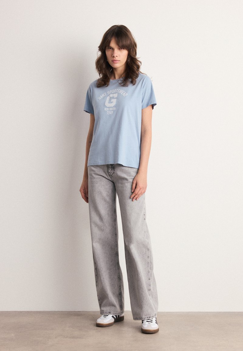 Ljusblå kortärmad T-shirt med tryckt logotyp, kombinerad med höga grå jeans och vita sneakers. Enfärgad bakgrund.