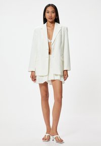 Witte blazer met een diepe V-hals, cropped fit en twee voorkruiszakken, gecombineerd met een bijpassende minirok en witte flip-flop hakken.